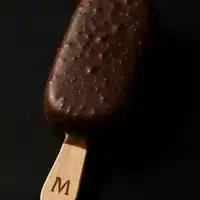 Magnum Classique : Plaisir Glacé au Chocolat Craquant Plaisir glacé : bâtonnet Magnum enrobé de chocolat craquant, tenu par un manche en bois marqué d'un 'M', sur fond sombre.