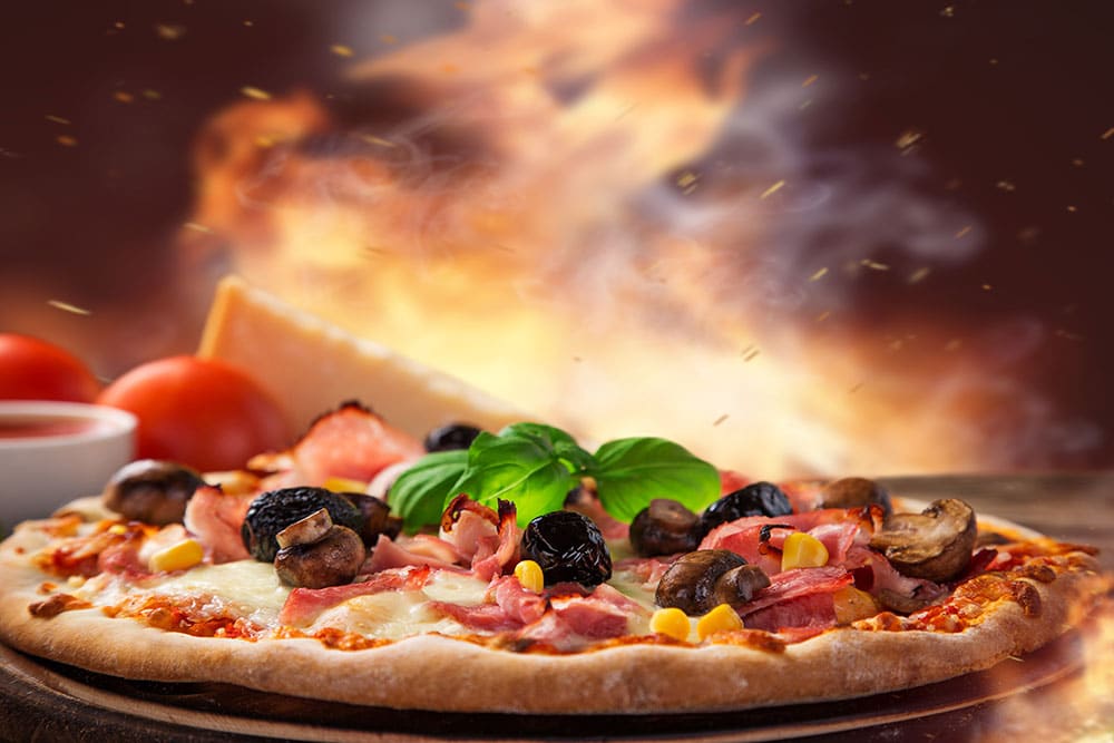 Pizza fraîche garnie de champignons, olives noires, jambon et basilic, sur fond de four à bois incandescent, avec parmesan et tomates.