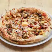 Pizza Champignons et Œuf : Saveur Rustique et Croûte Dorée Pizza appétissante à croûte épaisse, garnie de champignons, de morceaux de viande et d'un œuf au centre, servie sur une assiette blanche.