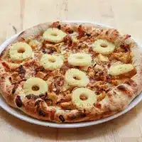 Délicieuse Pizza Hawaïenne Poulet Ananas Fraîche Pizza hawaïenne cuite au four sur plat blanc, garnie d'ananas en rondelles, poulet et fromage fondu, croûte dorée et croustillante.