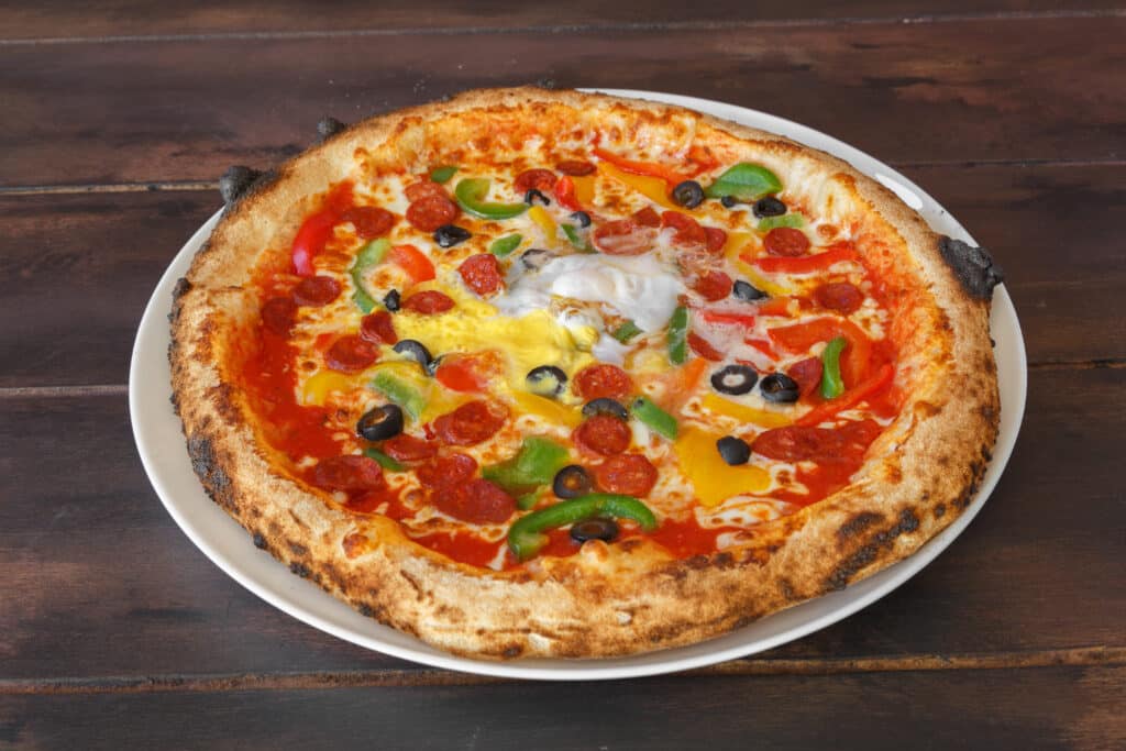 Pizza Napolitaine Pepperoni, Œuf et Légumes sur Bois Pizza napolitaine sur assiette blanche, garnie de pepperoni, poivrons colorés, olives noires et un œuf au centre, sur fond de bois sombre.