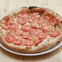 Pizza Tomate Fromage Artisanale Croustillante au Four Pizza artisanale cuite au four avec une croûte épaisse et dorée, généreusement garnie de tranches de tomate fraîche et de fromage fondu, présentée sur une assiette blanche.