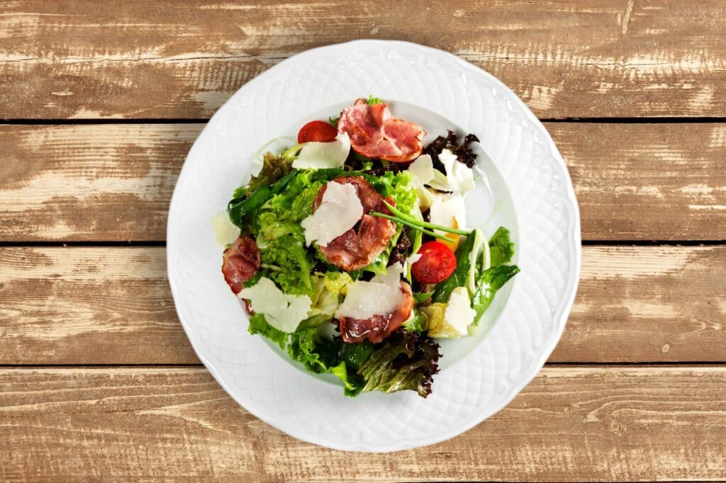 Salade Bacon Parmesan : Fraîcheur sur Fond Bois Salade fraîche avec lardons croustillants, copeaux de parmesan et tomates cerises, servie sur une assiette blanche sur fond de bois rustique.