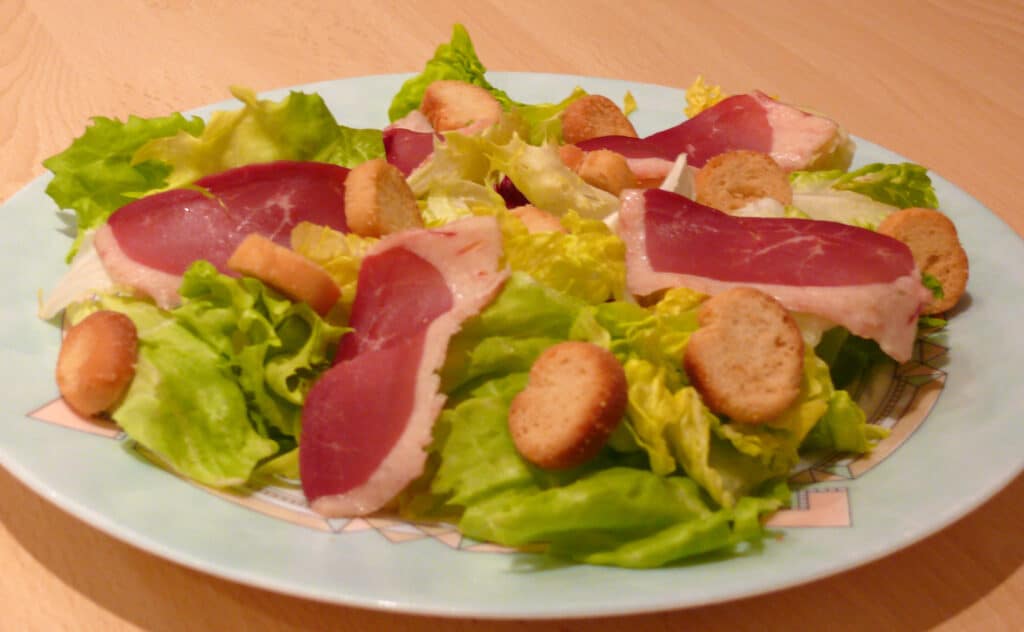 Salade Magret de Canard et Croûtons Frais Salade composée de laitue verte et jaune, garnie de fines tranches de magret de canard séché et de croûtons ronds, servie sur une assiette claire.