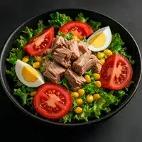 Salade de thon fraîche : Recette protéinée et légère Salade de thon copieuse avec morceaux de thon, tomates en tranches, œufs durs, maïs et laitue verte dans un bol noir.